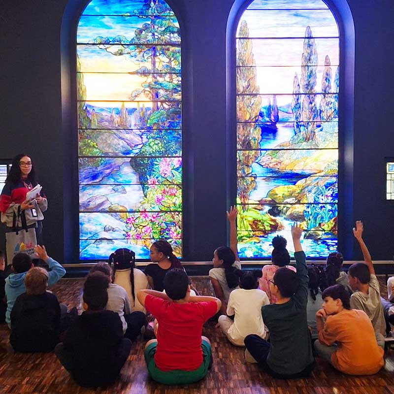 young kids learning about art, while seated infront of the Tiffany stained glass widows